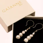 Paire de boucles d'oreilles pendantes Luna en perles de culture blanches et or laminé, présentée dans une boîte cadeau GAÏANNE beige à intérieur noir.