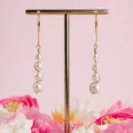 Boucles d'oreilles pendantes Luna en or laminé 14 carats et perles de culture blanches, suspendues à un présentoir doré parmi des fleurs roses.