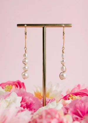 Boucles d'oreilles pendantes Luna en or laminé 14 carats et perles de culture blanches, suspendues à un présentoir doré parmi des fleurs roses.