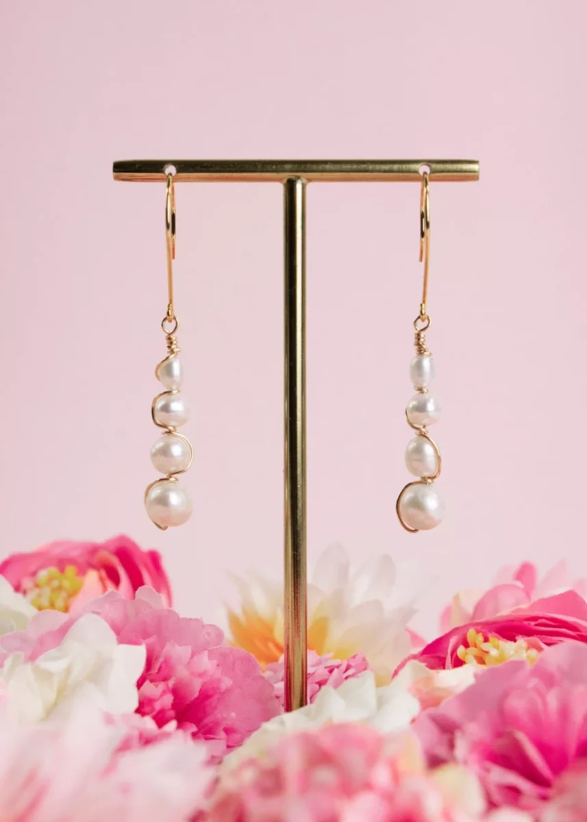 Boucles d'oreilles pendantes Luna en or laminé 14 carats et perles de culture blanches, suspendues à un présentoir doré parmi des fleurs roses.
