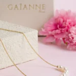 Collier Céleste en or laminé 14 carats et perles de culture d'eau douce blanches, présenté sur un support clair avec des fleurs roses.
