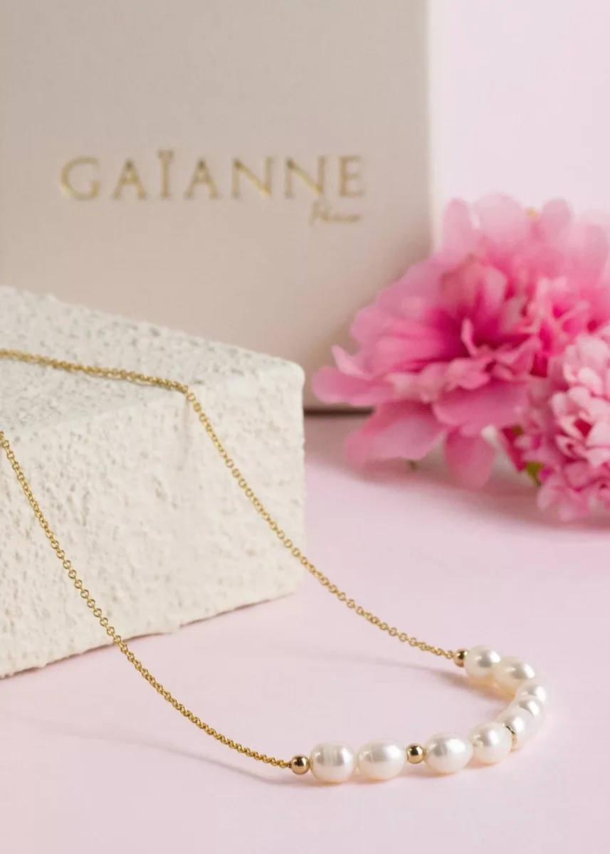 Collier Céleste en or laminé 14 carats et perles de culture d'eau douce blanches, présenté sur un support clair avec des fleurs roses.