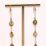 Boucles d'oreilles Nathéa de 8 cm suspendues à un présentoir doré, avec perles de Bohême aux reflets ambrés et chaînes fines en or laminé 14 carats.