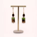 Vue d'ensemble des boucles d'oreilles Sandria de 6,5 cm, suspendues à un présentoir doré sur socle clair, avec perles vertes et brunes et attaches en or laminé.