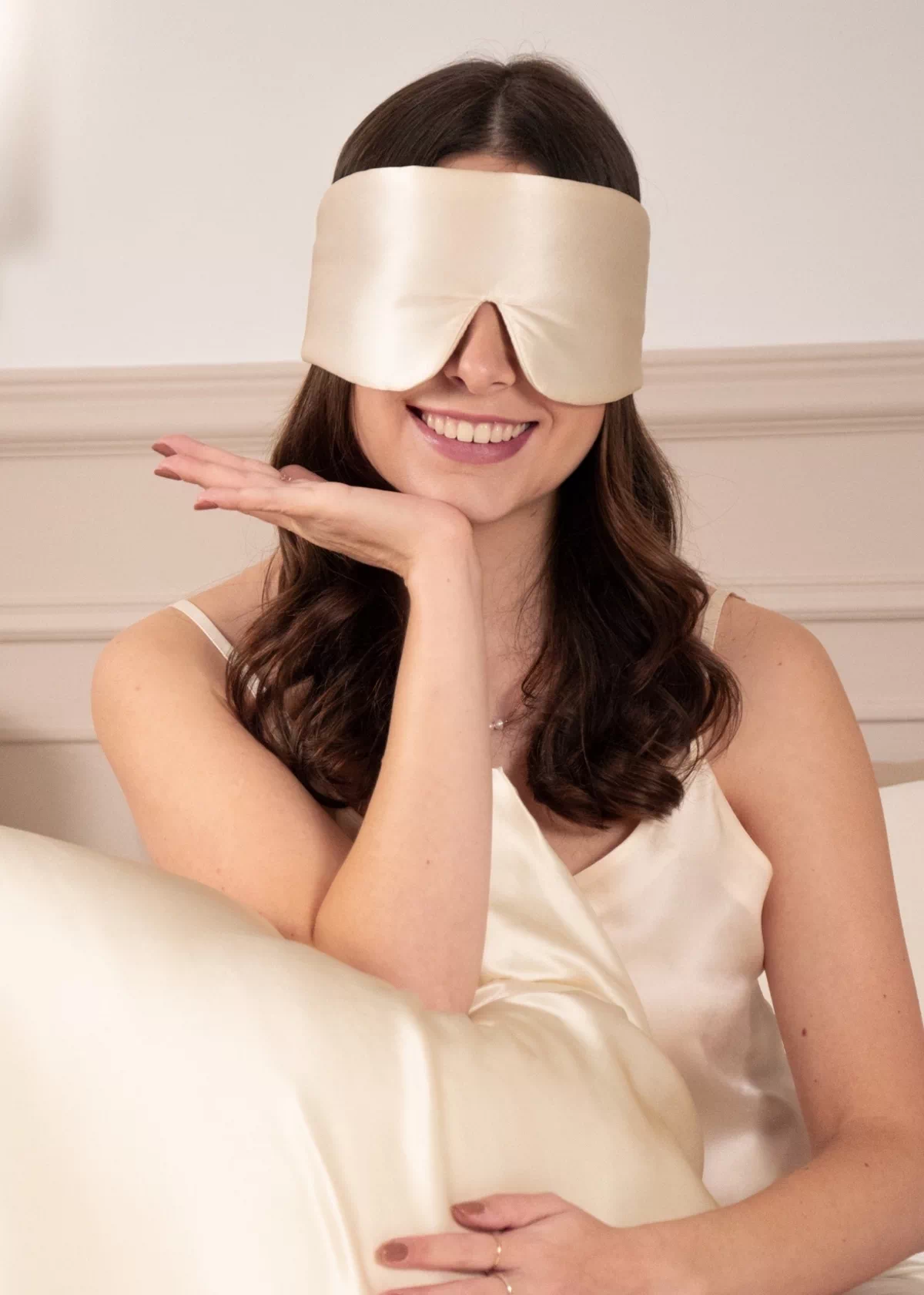 Femme souriante portant un masque de nuit bandeau réglable en soie Champagne devant un oreiller en soie clair.