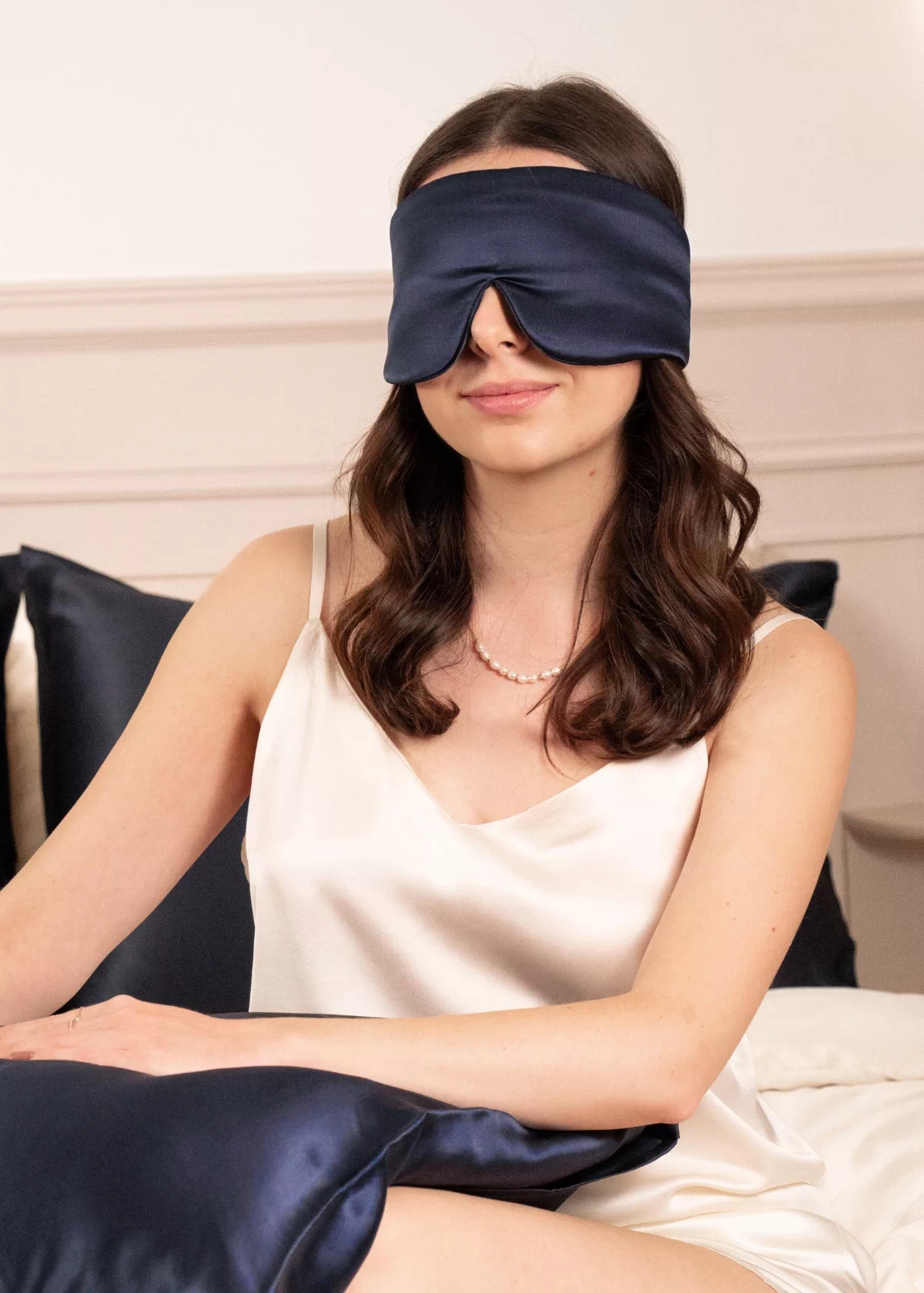 Femme portant un masque bandeau réglable en soie Bleu Nuit sur les yeux, assise sur un lit avec un oreiller assorti.