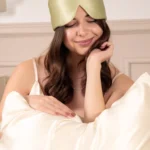 Femme portant un masque bandeau réglable en soie Vert Amande sur le front, la main posée sur le visage et un oreiller en soie devant elle.