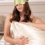 Femme portant un masque de nuit en soie Vert Amande sur les yeux, assise devant un oreiller en soie Champagne.