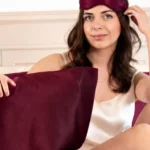 Femme ajustant un masque de nuit en soie Bordeaux sur le front, avec une taie d'oreiller en soie Bordeaux devant elle.