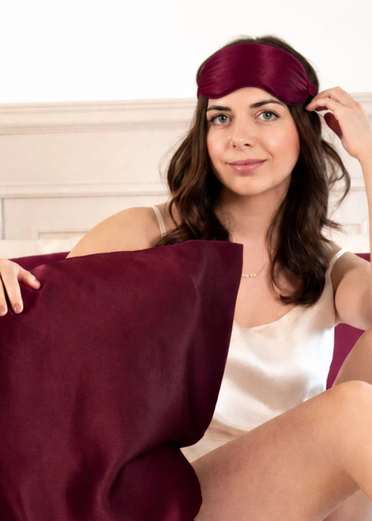 Femme ajustant un masque de nuit en soie Bordeaux sur le front, avec une taie d'oreiller en soie Bordeaux devant elle.