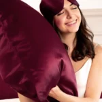 Femme souriante portant un masque de sommeil en soie Bordeaux sur le front et serrant une taie d'oreiller assortie contre elle.
