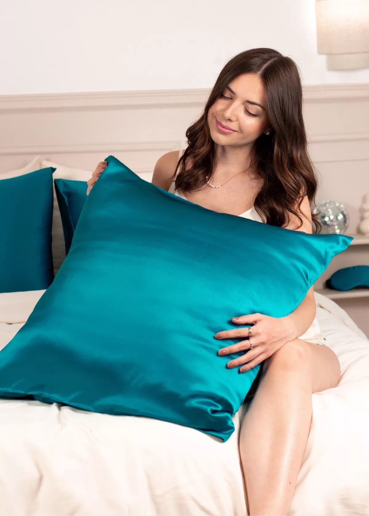 Femme assise sur un lit, caressant un oreiller recouvert d'une taie en soie Bleu Canard.